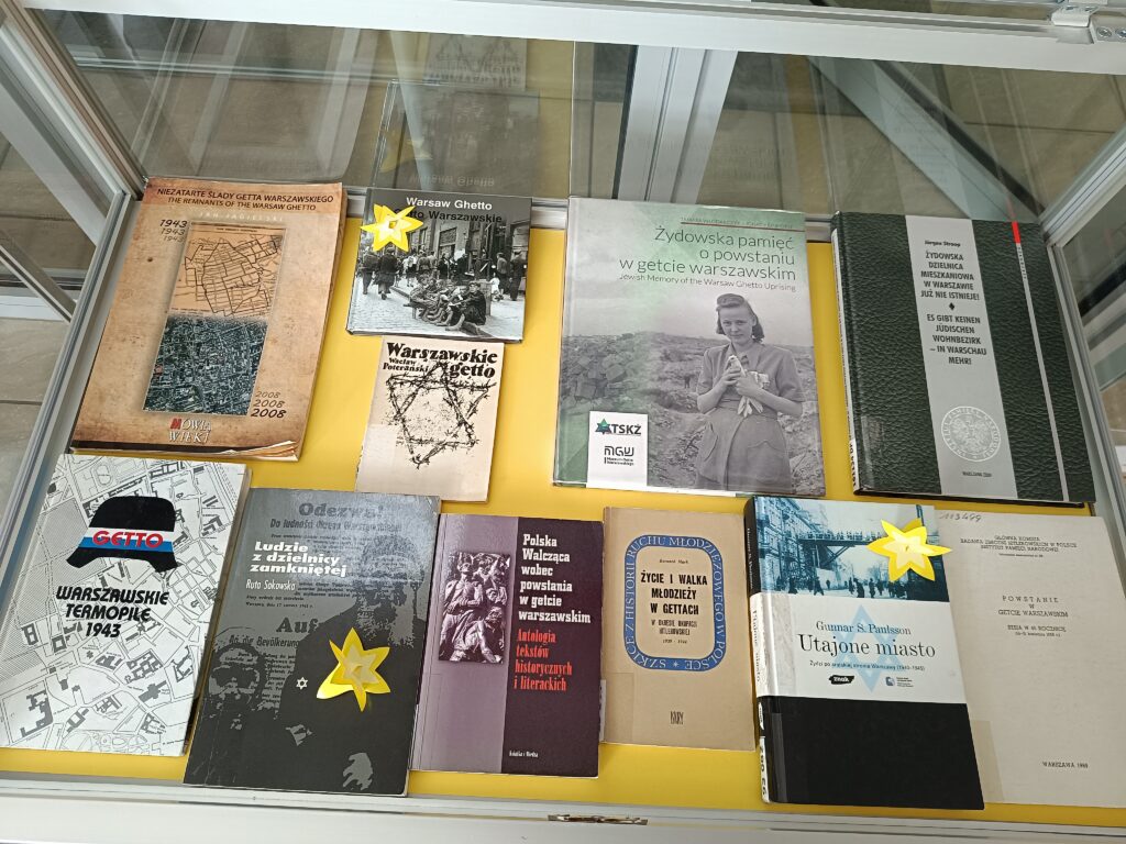 Widok z góry na szklaną gablotę z wystawą książek i publikacji historycznych dotyczących getta warszawskiego. Publikacje są ułożone na żółtym tle. Wśród nich znajdują się m.in. albumy ze zdjęciami, mapy getta oraz opracowania naukowe. Na kilku książkach przypięte są papierowe, żółte żonkile – symbol pamięci o powstaniu w getcie warszawskim. Widoczne tytuły to m.in. „Niezatarte ślady getta warszawskiego”, „Utajone miasto” oraz „Warszawskie Termopile 1943”.