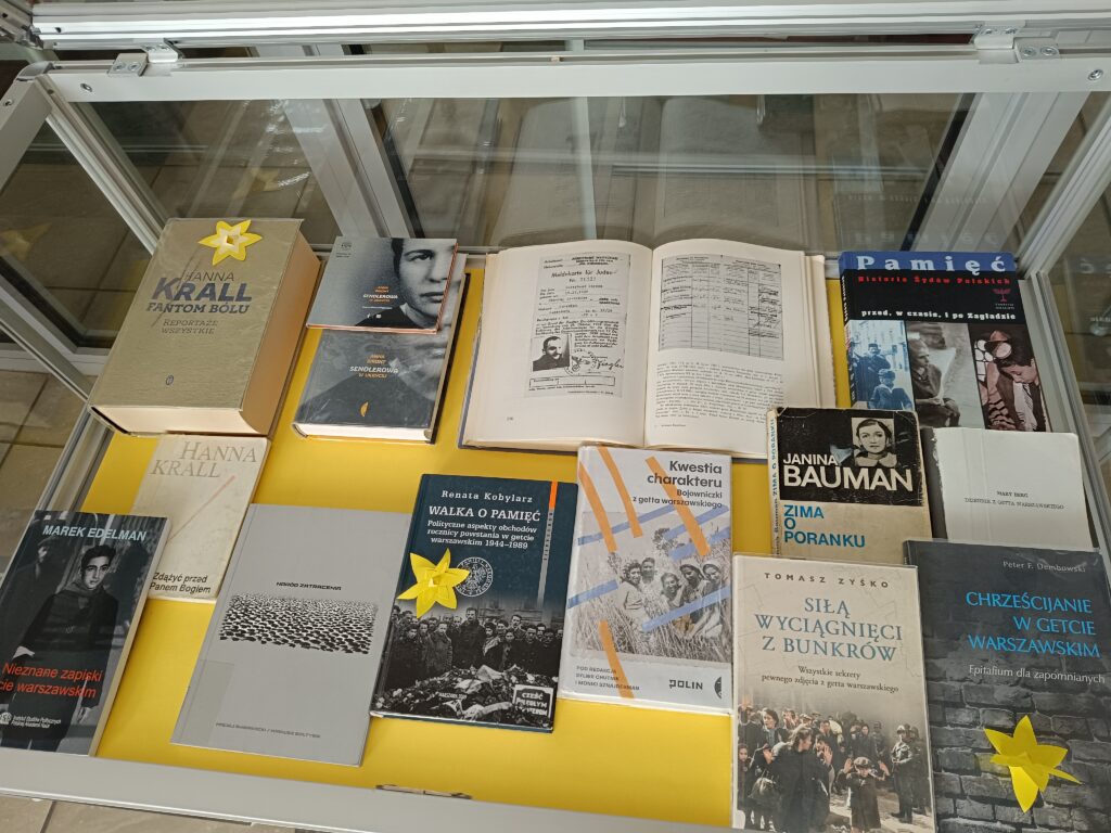 Zbliżenie na drugą część ekspozycji książkowej w szklanej gablocie. Na żółtym podłożu prezentowane są książki poświęcone osobistym świadectwom i analizom historycznym Zagłady. W centrum leży otwarta księga z dokumentami i fotografią portretową. Wokół niej znajdują się publikacje znanych autorów, m.in. „Fantom bólu” i „Zdążyć przed Panem Bogiem” Hanny Krall, „Zima o poranku” Janiny Bauman oraz „Nieznane zapiski z getta warszawskiego” Marka Edelmana. Ponownie pojawiają się żółte, papierowe żonkile przypięte do wybranych okładek.